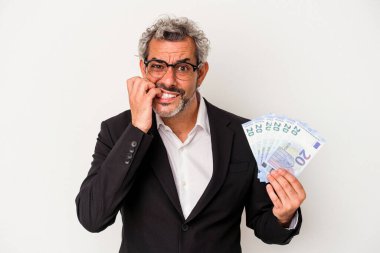 Elinde banknotlar tutan orta yaşlı bir iş adamı mavi arka planda izole edilmiş tırnak yiyen, gergin ve çok endişeli..