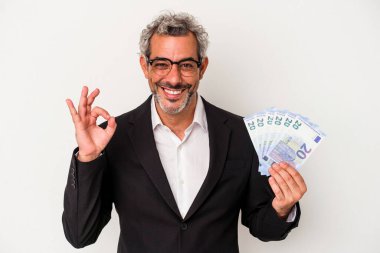 Orta yaşlı bir iş adamı mavi arka planda izole edilmiş faturaları tutuyor neşeli ve kendine güvenen iyi bir jest yapıyor..