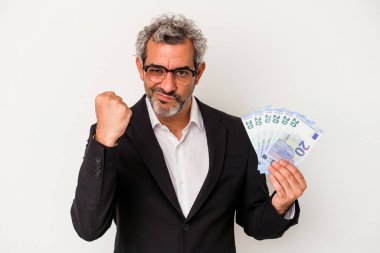 Elinde banknotlar tutan orta yaşlı bir iş adamı mavi arka planda yumruğunu kameraya gösteriyor, agresif bir yüz ifadesi var..