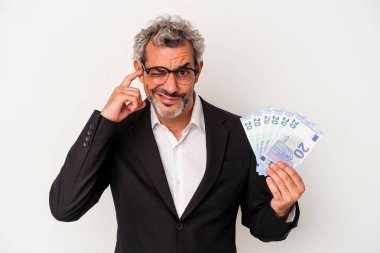 Elinde banknotlar tutan orta yaşlı bir iş adamı mavi arka planda, kulakları elleriyle kapatıyor..