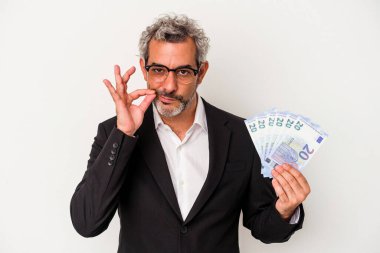 Orta yaşlı iş adamı mavi arka planda izole edilmiş banknotlar tutuyor ve parmakları dudaklarında bir sır saklıyor..