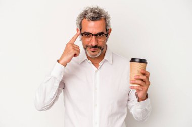 Orta yaşlı bir iş adamı beyaz arka planda izole edilmiş kahve tutuyor. Şakağı işaret ediyor, düşünüyor, bir göreve odaklanıyor..