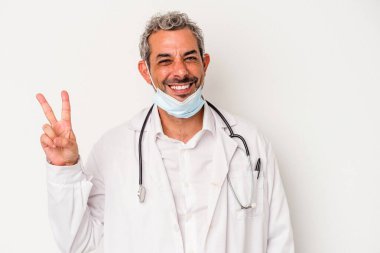 Virüs maskesi takan orta yaşlı bir doktor beyaz arka planda izole edilmiş neşeli ve kaygısız parmaklarıyla barış sembolü gösteriyor..