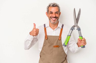 Orta yaşlı bahçıvan elinde makasla beyaz arka planda izole edilmiş gülümsüyor ve başparmağını kaldırıyor.