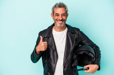 Mavi arka planda kask takan orta yaşlı motorcu beyaz adam gülümsüyor ve başparmağını kaldırıyor.