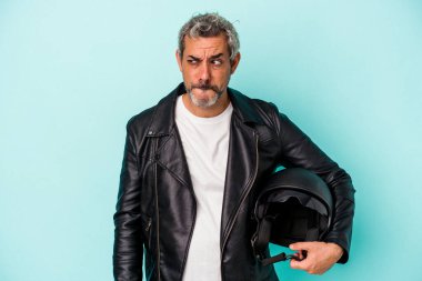 Mavi arka planda kask takan orta yaşlı motorcu beyaz adam şaşkın ve kararsız hissediyor..
