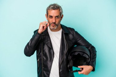 Beyaz kasklı orta yaşlı motosikletli adam mavi arka planda izole edilmiş dudaklarında parmakları olan bir sır saklıyor..