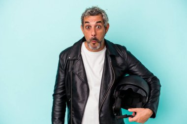 Mavi arka planda izole edilmiş miğfer tutan orta yaşlı motorcu beyaz adam omuzlarını silkeliyor ve gözleri açık..