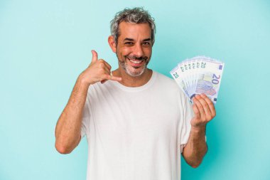 Elinde banknotlar tutan orta yaşlı beyaz bir adam mavi arka planda, parmakları olan bir cep telefonu hareketi gösteriyor..