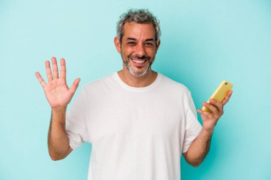 Elinde cep telefonu olan orta yaşlı beyaz bir adam mavi arka planda izole edilmiş, gülümseyen beş numarayı parmaklarıyla gösteriyor..
