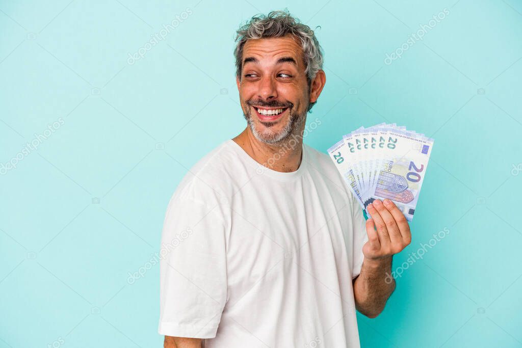 Middle age caucasian man holding bills isolated on blue background  looks aside smiling, cheerful and pleasant.