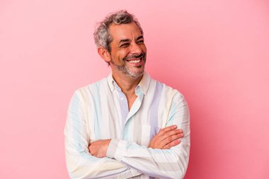 Pembe arka planda izole edilmiş orta yaşlı beyaz bir adam kollarını kavuşturarak gülümsüyor..