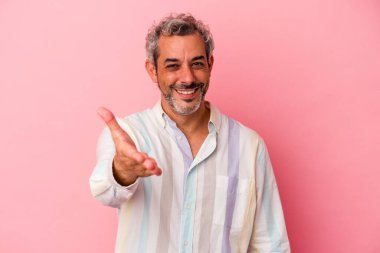 Orta yaşlı beyaz bir adam pembe arka planda izole edilmiş. Kameraya elini uzatıp selam veriyor..