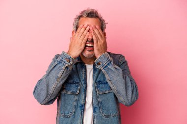 Pembe arka planda soyutlanmış orta yaşlı beyaz bir adam elleriyle gözlerini kapatıyor, büyük bir gülümsemeyle bir sürpriz bekliyor..