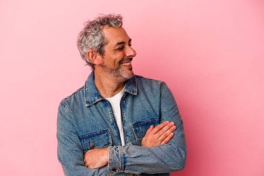 Pembe arka planda izole edilmiş orta yaşlı beyaz bir adam kollarını kavuşturarak gülümsüyor..