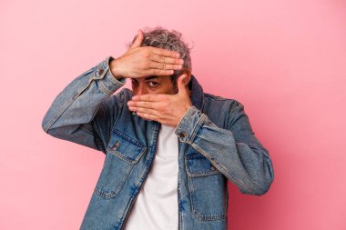 Orta yaşlı beyaz bir adam pembe arka planda izole edilmiş parmaklarıyla kameraya göz kırpıyor, yüzünü kapatmaktan utanıyor..