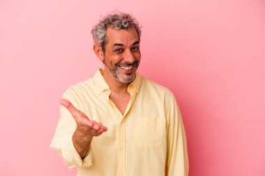 Orta yaşlı beyaz bir adam pembe arka planda izole edilmiş. Kameraya elini uzatıp selam veriyor..