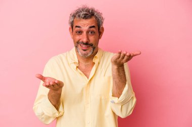 Pembe arka planda izole edilmiş orta yaşlı beyaz bir adam kollarıyla ölçek oluşturuyor, mutlu ve kendinden emin hissediyor..