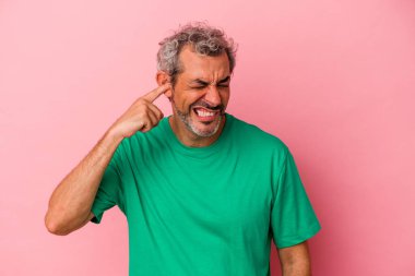 Orta yaşlı beyaz bir adam pembe arka planda izole edilmiş parmaklarıyla kulaklarını kapatıyor, gürültülü bir ortam yüzünden stresli ve çaresiz..