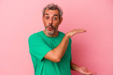 Orta yaşlı beyaz bir adam pembe arka planda izole edilmiş. Elleri arasında bir kopya alanı tutarken şok olmuş ve hayrete düşmüş..