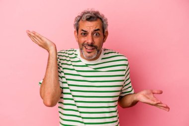 Orta yaşlı beyaz bir adam pembe arka planda izole edilmiş. Kafası karışmış ve omuz silkiyor..