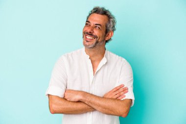 Mavi arka planda izole edilmiş orta yaşlı beyaz adam kollarını kavuşturarak gülümsüyor..