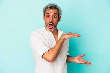 Orta yaşlı beyaz bir adam mavi arka planda izole edilmiş. Elleri arasında bir kopya alanı tutarken şok ve hayret içindeymiş..