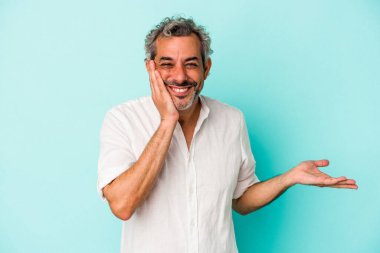 Mavi arka planda izole edilmiş orta yaşlı beyaz bir adam bir avuç içinde fotokopi alanı tutuyor, el üstünde tutuyor. Şaşırmış ve mutlu..