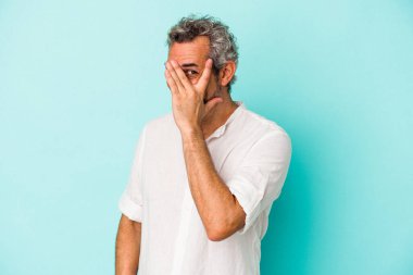Mavi arka planda izole edilmiş orta yaşlı beyaz bir adam kameraya parmaklarıyla göz kırpıyor, yüzünü kapatmaktan utanıyor..