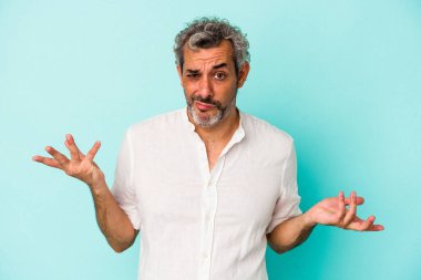 Orta yaşlı beyaz bir adam mavi arka planda izole edilmiş sorgulama sırasında omuz silkiyor ve şüphe duyuyor..