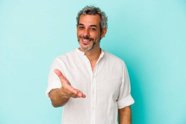 Mavi arka planda izole edilmiş orta yaşlı beyaz bir adam kameraya elini uzatarak selam veriyor..
