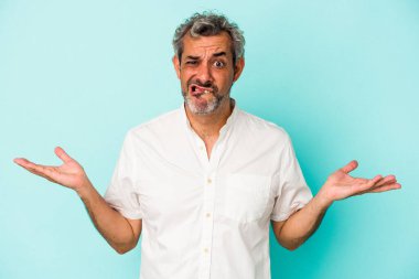 Orta yaşlı, beyaz bir adam mavi arka planda izole edilmiş. Kafası karışmış ve omuz silkiyor..