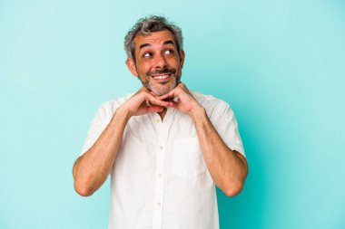 Mavi arka planda izole edilmiş orta yaşlı beyaz bir adam ellerini çenesinin altında tutuyor, mutlu bir şekilde yana bakıyor..