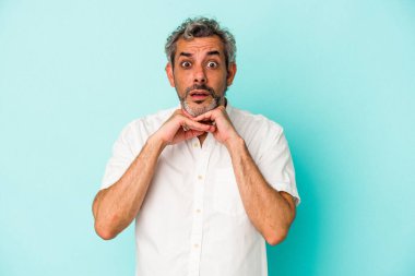 Orta yaşlı, beyaz bir adam mavi arka planda izole edilmiş şans diliyor, şaşırmış ve ağzını açıp önüne bakıyor..