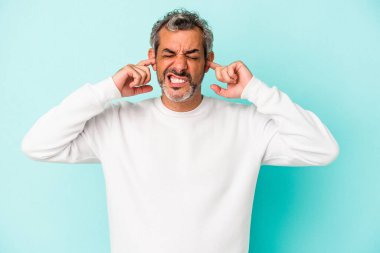 Orta yaşlı beyaz bir adam mavi arka planda izole edilmiş parmaklarıyla kulaklarını kapatıyor, gürültülü bir ortam yüzünden stresli ve çaresiz..