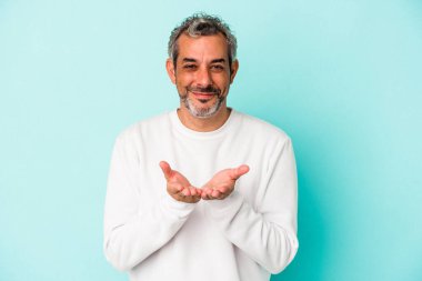 Orta yaşlı, beyaz bir adam mavi arka planda izole edilmiş. Elinde avuç içi olan bir şeyle kameraya sunuyor..
