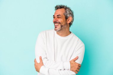 Mavi arka planda izole edilmiş orta yaşlı beyaz adam kollarını kavuşturarak gülümsüyor..