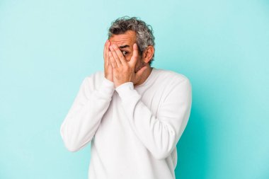 Orta yaşlı, beyaz bir adam mavi arka planda izole edilmiş. Korkmuş ve gergin parmaklarıyla göz kırp..