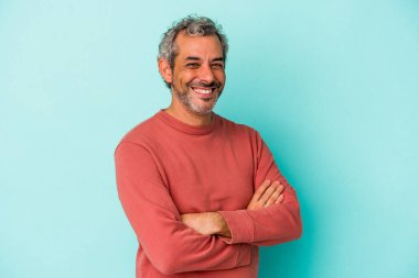 Mavi arka planda izole edilmiş orta yaşlı beyaz bir adam. Kendine güveniyor, kararlılıkla kollarını kavuşturuyor..