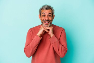 Orta yaşlı, beyaz bir adam mavi arka planda izole edilmiş şans diliyor, şaşırmış ve ağzını açıp önüne bakıyor..