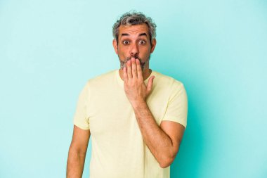 Mavi arka planda izole edilmiş orta yaşlı beyaz adam şok olmuş, ağzı ellerle kaplanmış, yeni bir şey keşfetmeye hevesli..