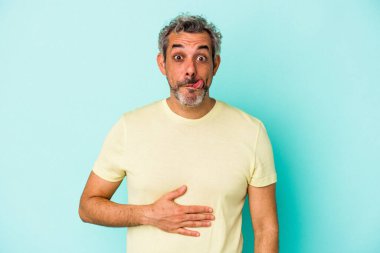 Mavi arka planda izole edilmiş orta yaşlı beyaz adam karnına dokunuyor, nazikçe gülümsüyor, yemek yiyor ve tatmin oluyor..