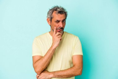 Mavi arka planda izole edilmiş orta yaşlı beyaz adam şüpheli ve şüpheci bir ifadeyle yana bakıyor..
