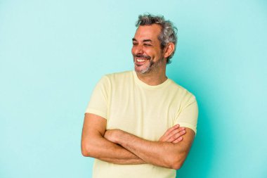 Mavi arka planda izole edilmiş orta yaşlı beyaz adam kollarını kavuşturarak gülümsüyor..
