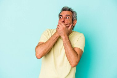 Orta yaşlı beyaz bir adam mavi arka planda izole edilmiş. Düşünceli bir şekilde bir fotokopi odasına bakıyor. Ağzını eliyle kapatıyor..