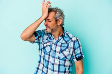 Orta yaşlı beyaz bir adam mavi arka planda izole edilmiş bir şekilde bir şeyi unutuyor. Alnına avuç içi ve kapalı gözlerle vuruyor..