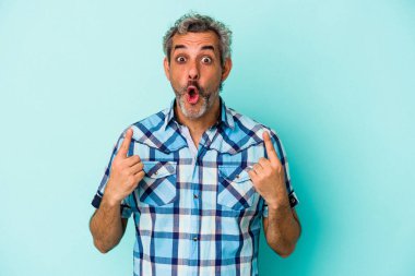 Mavi arka planda izole edilmiş orta yaşlı beyaz bir adam ağzı açık bir şekilde üst tarafı işaret ediyor..