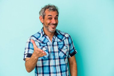 Mavi arka planda izole edilmiş orta yaşlı beyaz bir adam kameraya elini uzatarak selam veriyor..