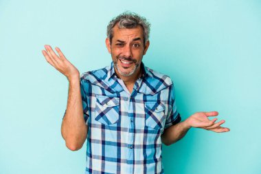 Orta yaşlı, beyaz bir adam mavi arka planda izole edilmiş. Kafası karışmış ve omuz silkiyor..
