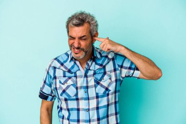 Orta yaşlı beyaz bir adam mavi arka planda izole edilmiş kulakları elleriyle kapatıyor..